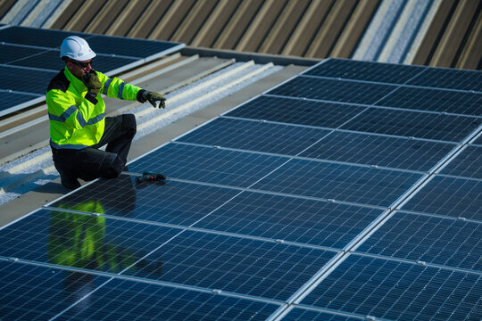 A photovoltaic technician wearing a safety vest and hard hat skilfully crouches in warm light calling in the inspection results on the sweeping metal roof of a manufacturing plant.