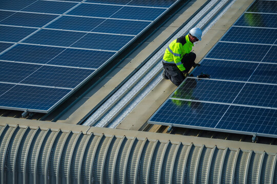 A renewable energy specialist in reflective workwear and helmet attentively crouches over the panels for a final evening test on a commercial factory rooftop photovoltaic construction site.