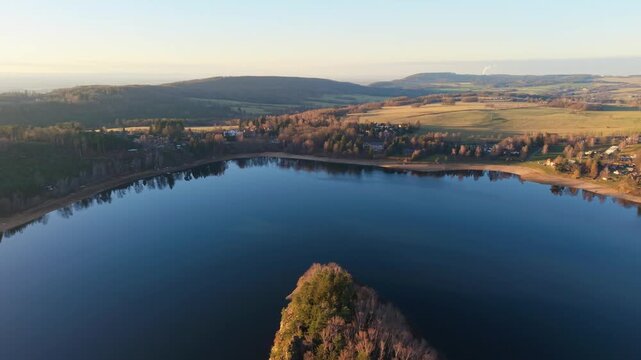 Aerial view of Sec Dam at sunset with a rocky peninsula extending into the calm water surrounded by rolling hills and forest in Czechia.