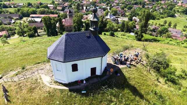 Aerial view of the Chapel of Saint Anne on a grassy hill overlooking the village of Vysker, surrounded by lush green forests and rolling hills in Vysker, Liberecky kraj, Czechia.
