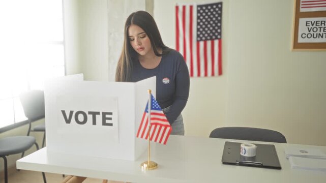 Woman places ballot into a voting booth inside a public building while wearing an i voted sticker and standing by a small american flag; patriotism civic duty.