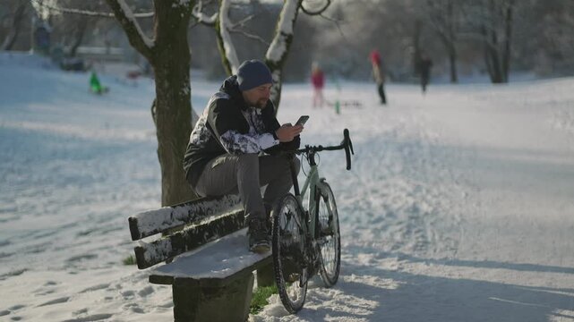 Man using a mobile phone for GPS during a winter bike commute in Munich. A cyclist stops on a snow-covered bench to plan his route using a digital device, showing a lifestyle of year-round cycling.