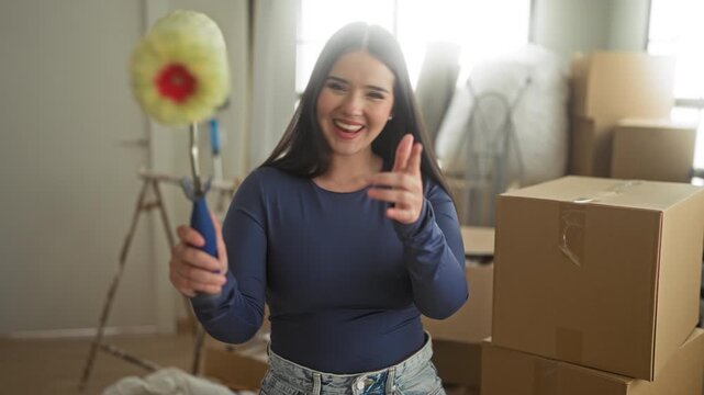 Woman holding paint roller and waving hand among cardboard moving boxes in building; joy fresh start.