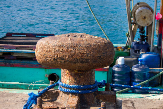 Rusty metal mooring bollard with a blue rope tied around it on a concrete pier, securing a fishing boat