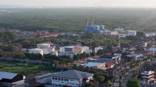 Aerial view of Pertamina Fuel Depot with large storage tanks, surrounding industrial buildings, lush greenery, and nearby road traffic in Kecamatan Denpasar Selatan, Bali, Indonesia.