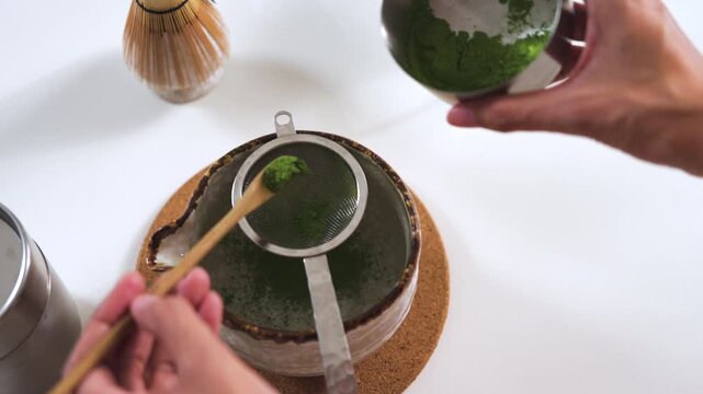 High-angle shot of hands using a bamboo scoop to sift organic matcha green tea powder through a metal strainer into a traditional ceramic bowl. Cafe beverage preparation concept.