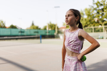 Fototapeta premium Young girl practices tennis on a sunny day at a local court focusing on her next move as she holds a tennis ball in hand ready to serve or rally