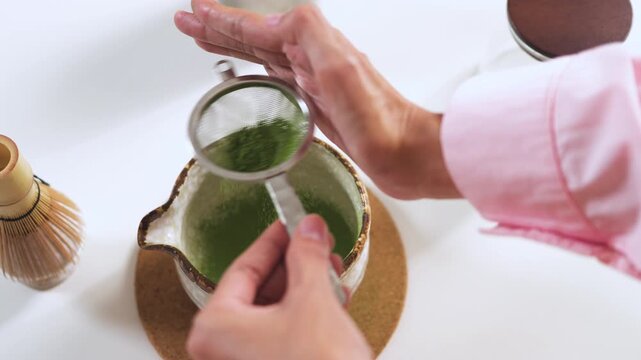 High-angle close-up of hands tapping a metal strainer to sift organic matcha green tea powder into a ceramic bowl. Traditional Japanese tea preparation and cafe drink concept.