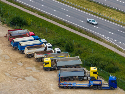 Aerial view of a row of parked dump trucks and a flatbed truck in a dirt lot adjacent to a highway, Vilnius, Vilniaus apskritis, Lithuania.