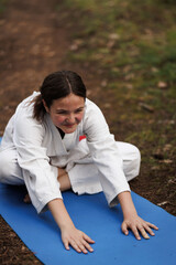 Young girl in white karate uniform stretching on a blue mat outdoors during martial arts training, smiling and focused while practicing flexibility and warm-up exercises in a park © Nikola Spasenoski