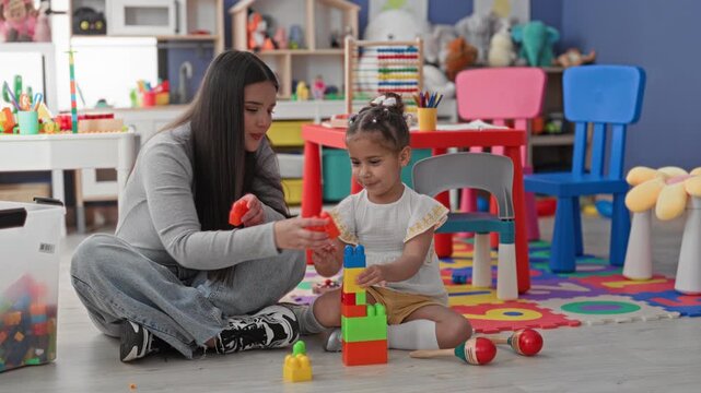 Woman teacher guides little girl stacking colorful toy blocks, placing a block on a tall tower while seated on the floor in playroom building; nurturing bond.