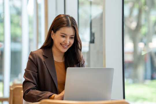 Smiling young woman using a laptop in a bright office, working at a table near a window.