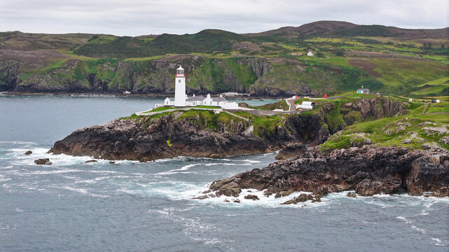 Aerial view of Fanad Head Lighthouse perched on a rugged cliffside overlooking the Atlantic Ocean, surrounded by green hills and rocky coastline in Baile Lair, County Donegal, Ireland.