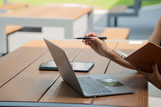 Close-up of a woman working at an outdoor table with a laptop, phone, notebook and pen, showing study, planning, remote work and modern business in a calm setting.