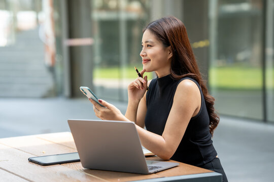 Young woman working outdoors with a laptop and smartphone, smiling while thinking at a table near a modern office building