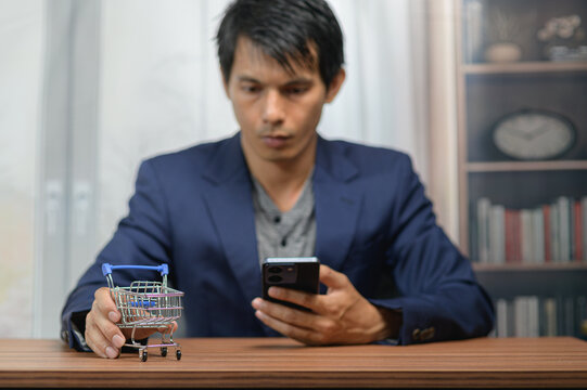 Man Shopping Online Using Smartphone with Small Toy Shopping Cart on Wooden Table
