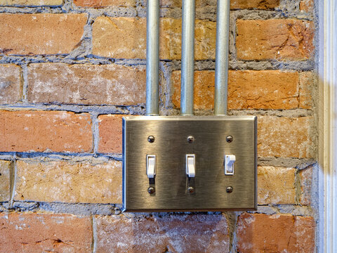 A switch plate with 3 switches and conduits on an interior brick wall