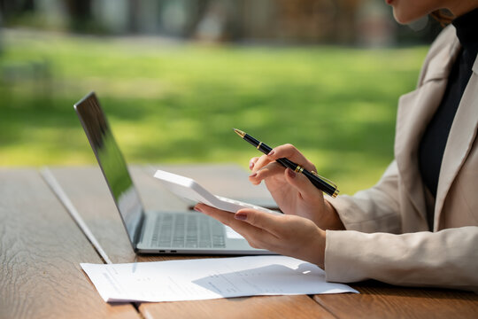 Person working on a laptop and taking notes outdoors at a table with papers and a pen