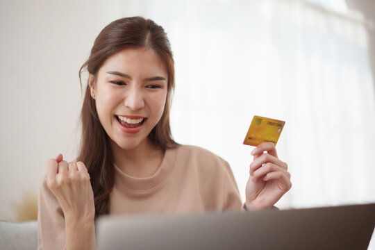 Happy Asian woman celebrating a successful online payment while holding a credit card and shopping on a laptop.