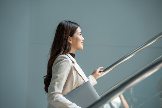 Smiling businesswoman in a light blazer walking up stairs with a laptop in a modern office building