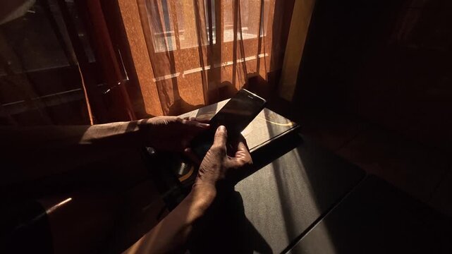 Gresik Indonesia - August 5, 2026: Side Angle Slow Motion Shot of Person Scrolling and Using Mobile Phone in Warm Window Sunlight