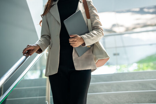 A businesswoman in a blazer climbs stairs while holding a laptop in a modern office building, with bright natural light and a clean corporate interior.