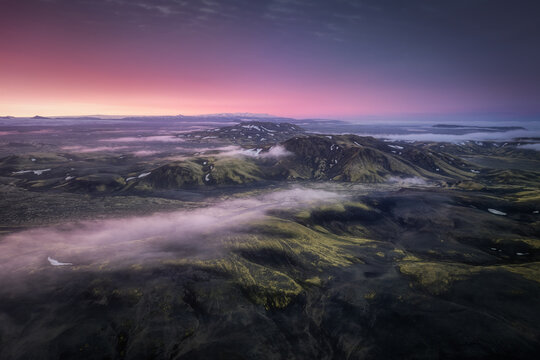 Aerial view of Laki volcanic landscape with moss-covered hills, dark volcanic rock, and low-hanging mist under a vibrant sunset sky in Skaftarhreppur, Iceland.