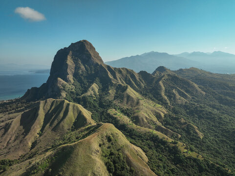 Aerial view of Nuca Molas island with its prominent rocky peak, lush green slopes, and surrounding blue sea under a clear sky in Satar Mese Barat, East Nusa Tenggara, Indonesia.