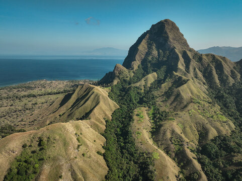 Aerial view of Nuca Molas island featuring rugged green hills and steep slopes meeting the blue sea in Satar Mese Barat, East Nusa Tenggara, Indonesia.