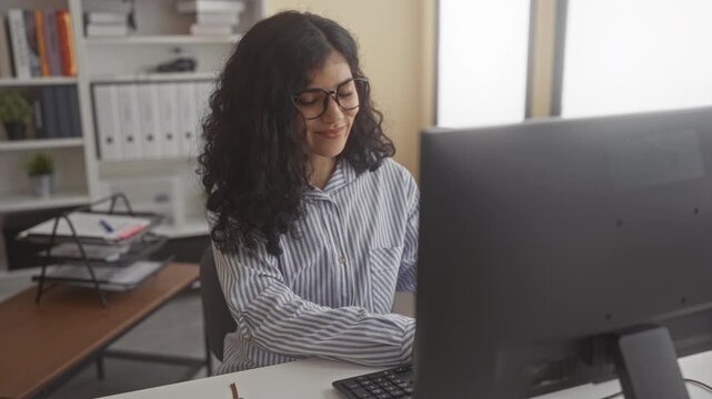 Young woman seated at desk types then forms heart with hands while smiling at computer in a building office; affection connection.