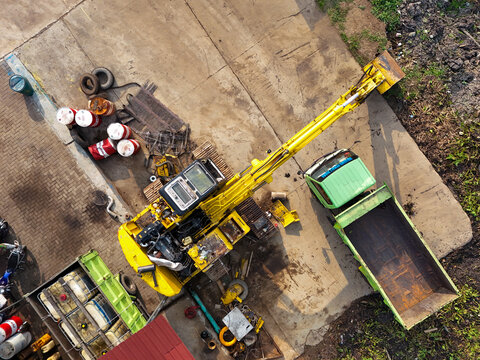 Aerial view of a yellow excavator undergoing maintenance with a green dump truck and oil drums on a concrete surface in Central Java, Indonesia.