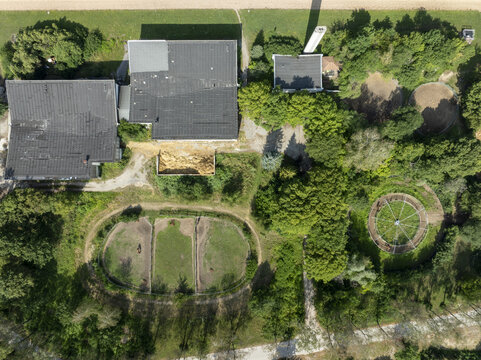 Aerial view of an equestrian facility with stables, horse paddocks, and a round pen surrounded by lush green trees and fields in Munich, Bavaria, Germany.
