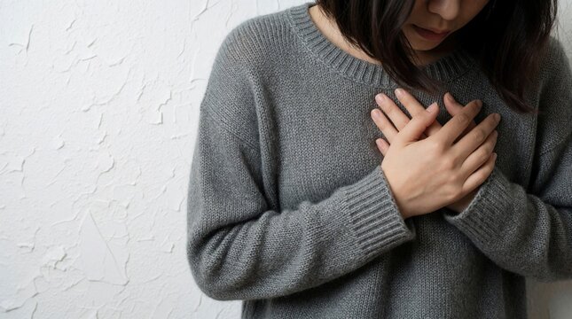Young woman in grey sweater touches chest with hands due to sharp heart pain. Person feels sudden discomfort anxiety respiratory trouble indoors. Concept of cardiovascular disease medical checkup