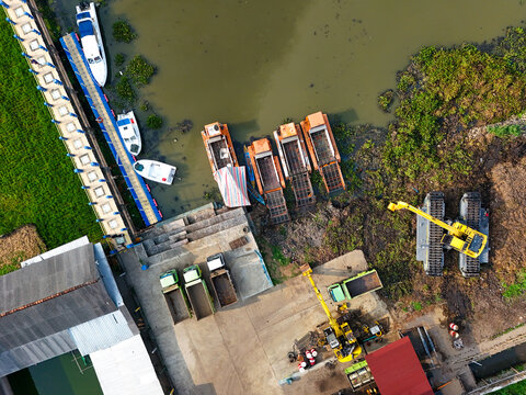 Aerial view of heavy-duty machinery and excavators removing sediment from Rawa Pening lake to maintain water depth, with trucks and boats docked nearby in Tuntang, Central Java, Indonesia.