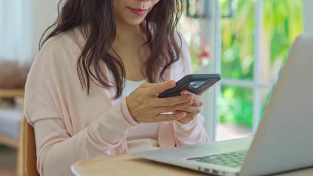 Asian female freelancer holding a mobile phone, messaging or checking notifications in front of a laptop