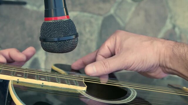POV (slow zoom in): man playing acoustic guitar in front of a microphone. A guitar player in front of a microphone. A guitarist strumming a guitar. An artist plucking a guitar. Musician playing guitar