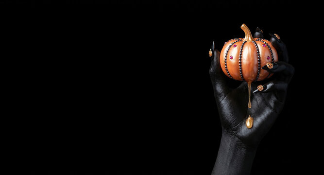 black hand with decorated nails holding a luxury golden pumpkin with dripping gold syrup on a black background representing glamorous Halloween concept, gothic luxury, and dark magical celebration.