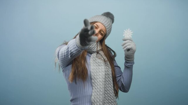 Woman wearing knit hat, scarf and gloves holding snowflake and reaches hand toward camera in studio; winter joy playful.