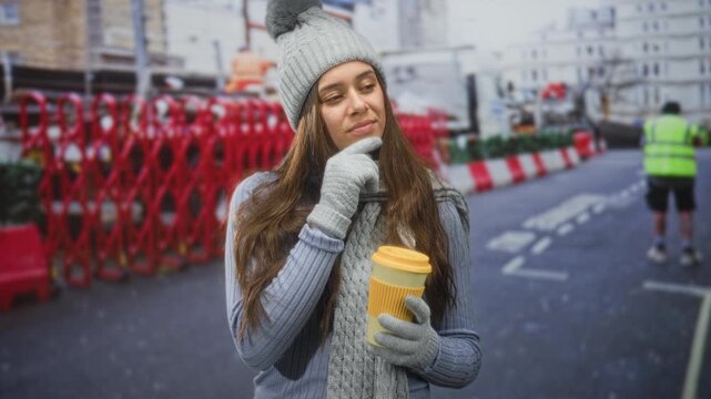 Woman holding a yellow takeaway coffee cup, finger to chin gesture and visible face, wearing winter beanie and gloves on a street; thoughtful warmth.