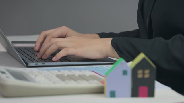 Typing on laptop near finance calculator and miniature house model to plan mortgage budget, achieving future home ownership goal successfully