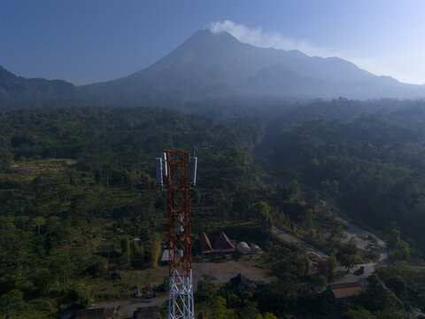 Aerial view of a telecommunications tower standing in a lush forest with the active Mount Merapi volcano in the background in Kecamatan Cangkringan, Daerah Istimewa Yogya, Indonesia.