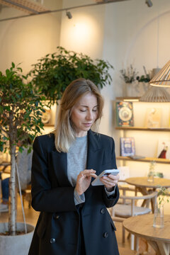 Calm successful businesswoman restaurant executive standing in dining space watching scrolling news on smartphone. Positive lady ceo cafe business owner texting or reading reviews on mobile phone.