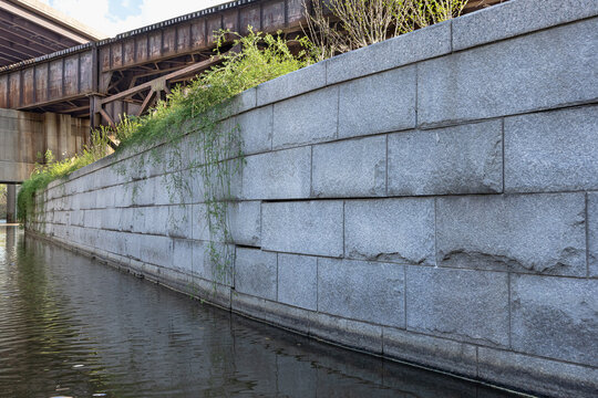 Stone wall of the historic Shockoe Bottom canal system in Richmond Virginia with plants growing over the side, and an oxidized iron railroad  trestle in the background.