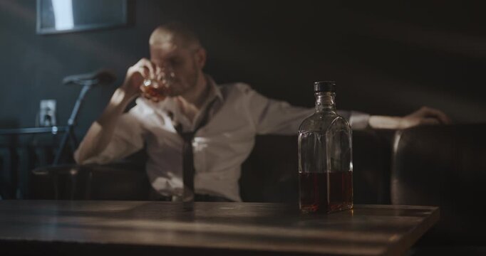 Bearded man in a loosened tie reaches for a whiskey glass while relaxing alone in a dim room.
