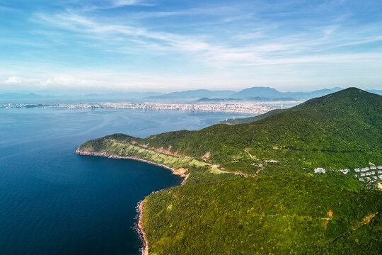 Dense green jungle foliage blankets mountain terrain extending to a peaceful blue ocean. A coastal city lines the horizon under a bright daytime sky