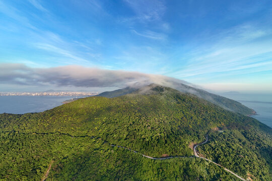 Lush green forest hills cover the Son Tra Peninsula landscape as white clouds drift over the peak near the coastline of Danang city during a calm morning