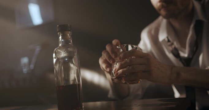 Close-up of a seated man holding a glass of whiskey in both hands beside a bottle on the table.