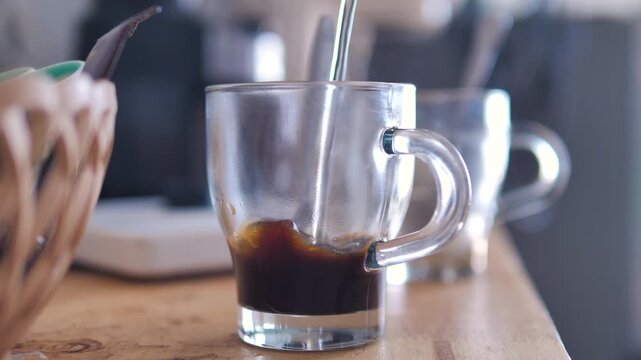 Close-up video of black coffee being brewed in a clear glass cup, with a coffee maker and cup in a cozy indoor setting