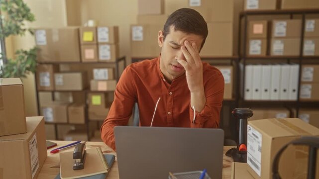 Man at laptop surrounded by shipping boxes rubbing forehead and holding glasses in a small business building; small business fatigue.