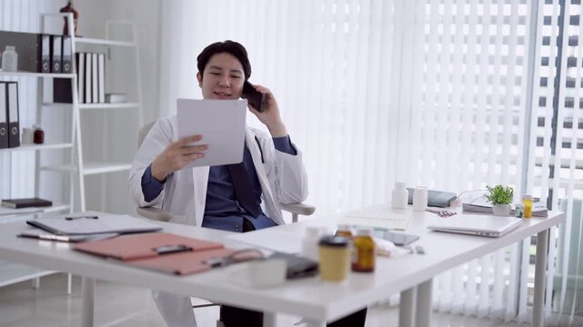 Young Asian male doctor reviewing medical report while taking phone call in bright modern office with copy space